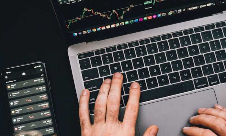 Hands typing on a laptop with cryptocurrency charts and smartphone display in view, focusing on digital finance.